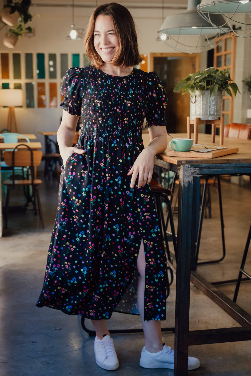 A woman smiling and posing in a midi dress with a black and rainbow polka dot print, featuring a shirred bodice and elasticated puff sleeves.