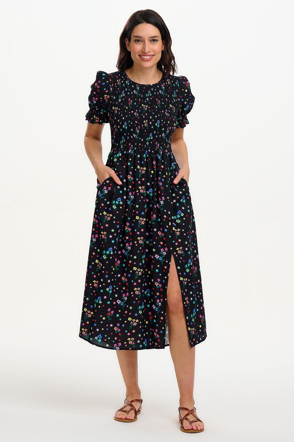 A model wearing a black midi dress with a rainbow sprigs print, featuring colorful ditsy flowers and stars, with a shirred bodice, elasticated puff sleeves, and a split hem.