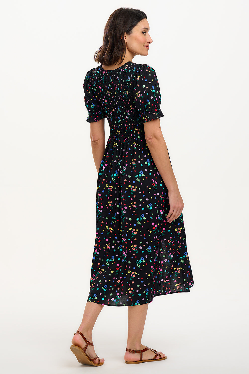 A model wearing a black midi dress with a rainbow sprigs print, featuring colorful ditsy flowers and stars, with a shirred bodice, elasticated puff sleeves, and a split hem.