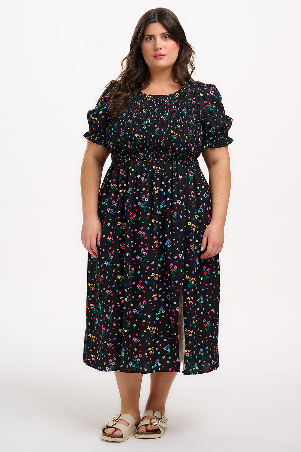 A model wearing a black midi dress with a rainbow sprigs print, featuring colorful ditsy flowers and stars, with a shirred bodice, elasticated puff sleeves, and a split hem.
