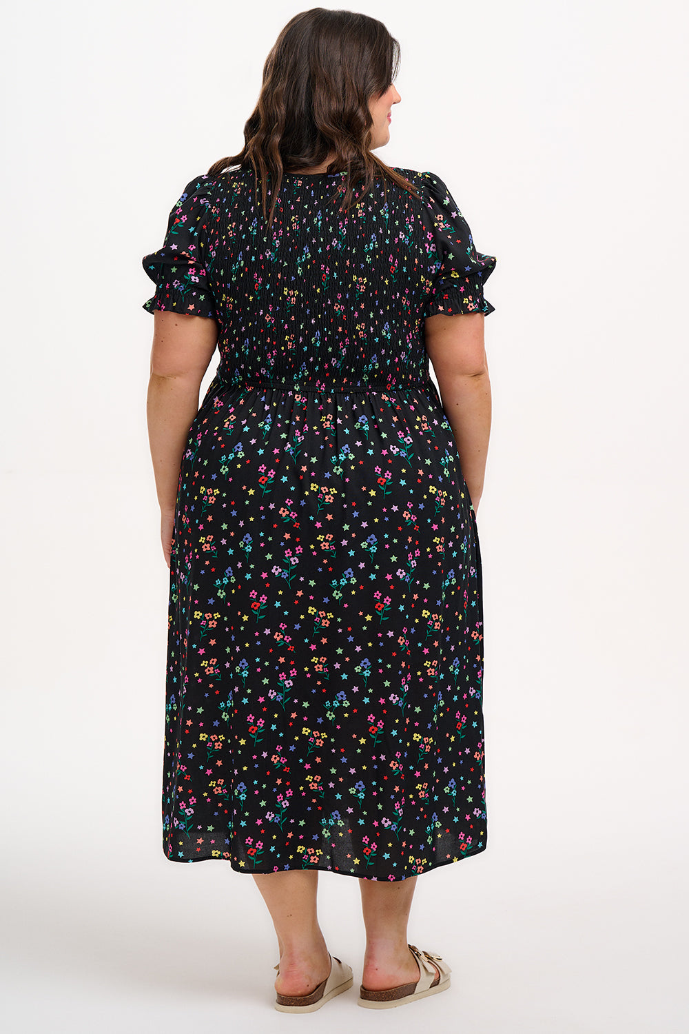 A model wearing a black midi dress with a rainbow sprigs print, featuring colorful ditsy flowers and stars, with a shirred bodice, elasticated puff sleeves, and a split hem.