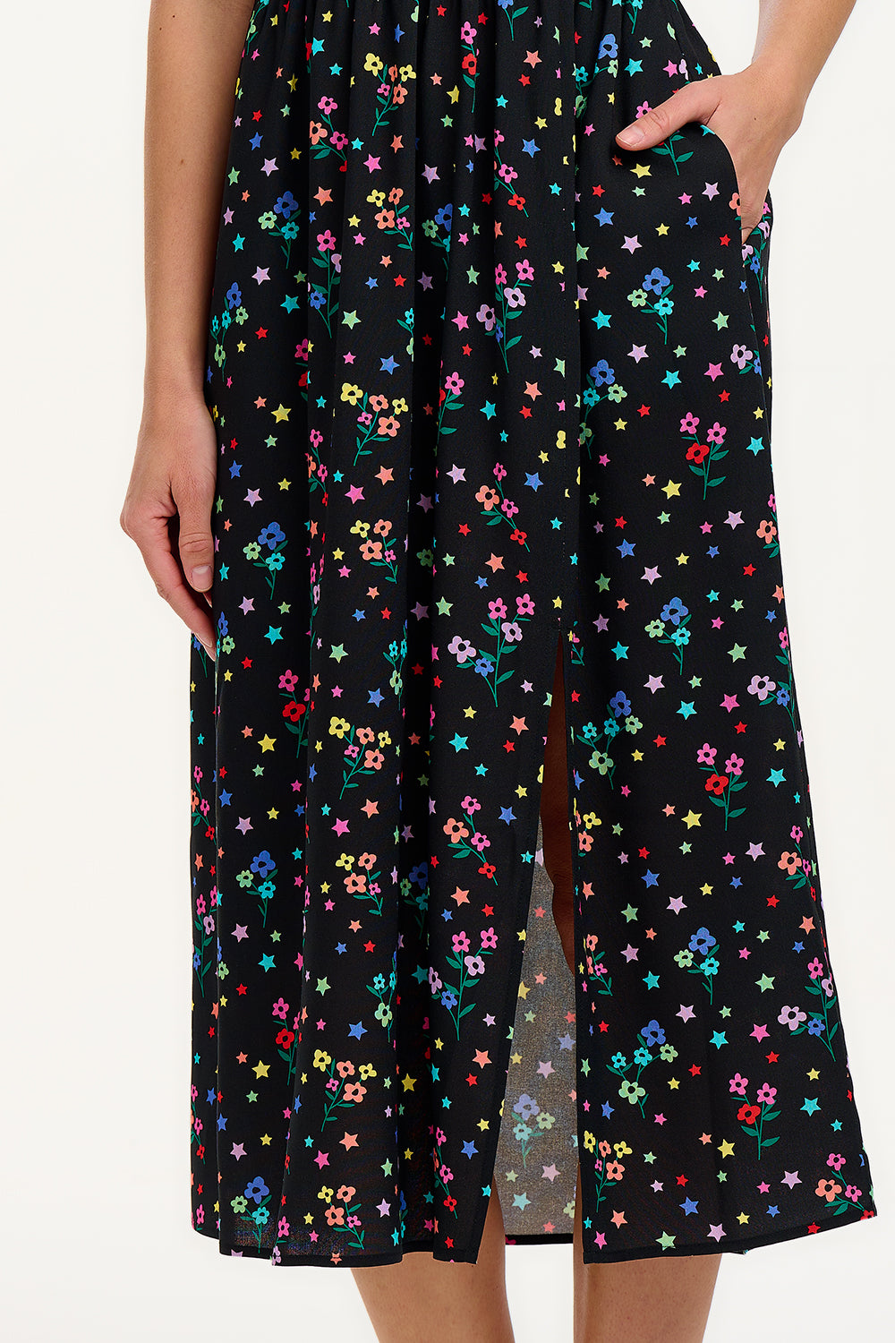 A model wearing a black midi dress with a rainbow sprigs print, featuring colorful ditsy flowers and stars, with a shirred bodice, elasticated puff sleeves, and a split hem.