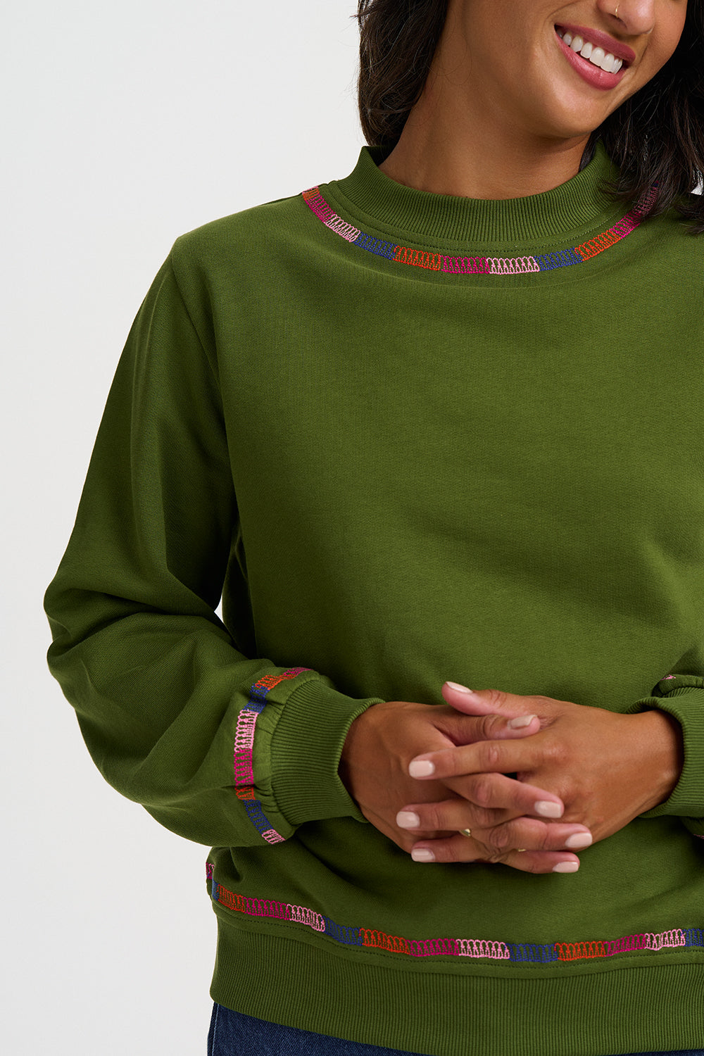 A close-up of a woman wearing an olive green crewneck sweatshirt with colorful geometric embroidered trim around the collar, cuffs, and hem.