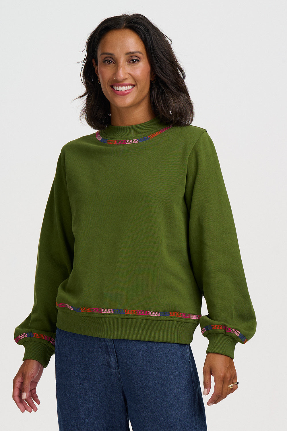 A woman wearing an olive green crewneck sweatshirt with patterned trim at the collar and cuffs, paired with blue jeans, against a light gray studio background.