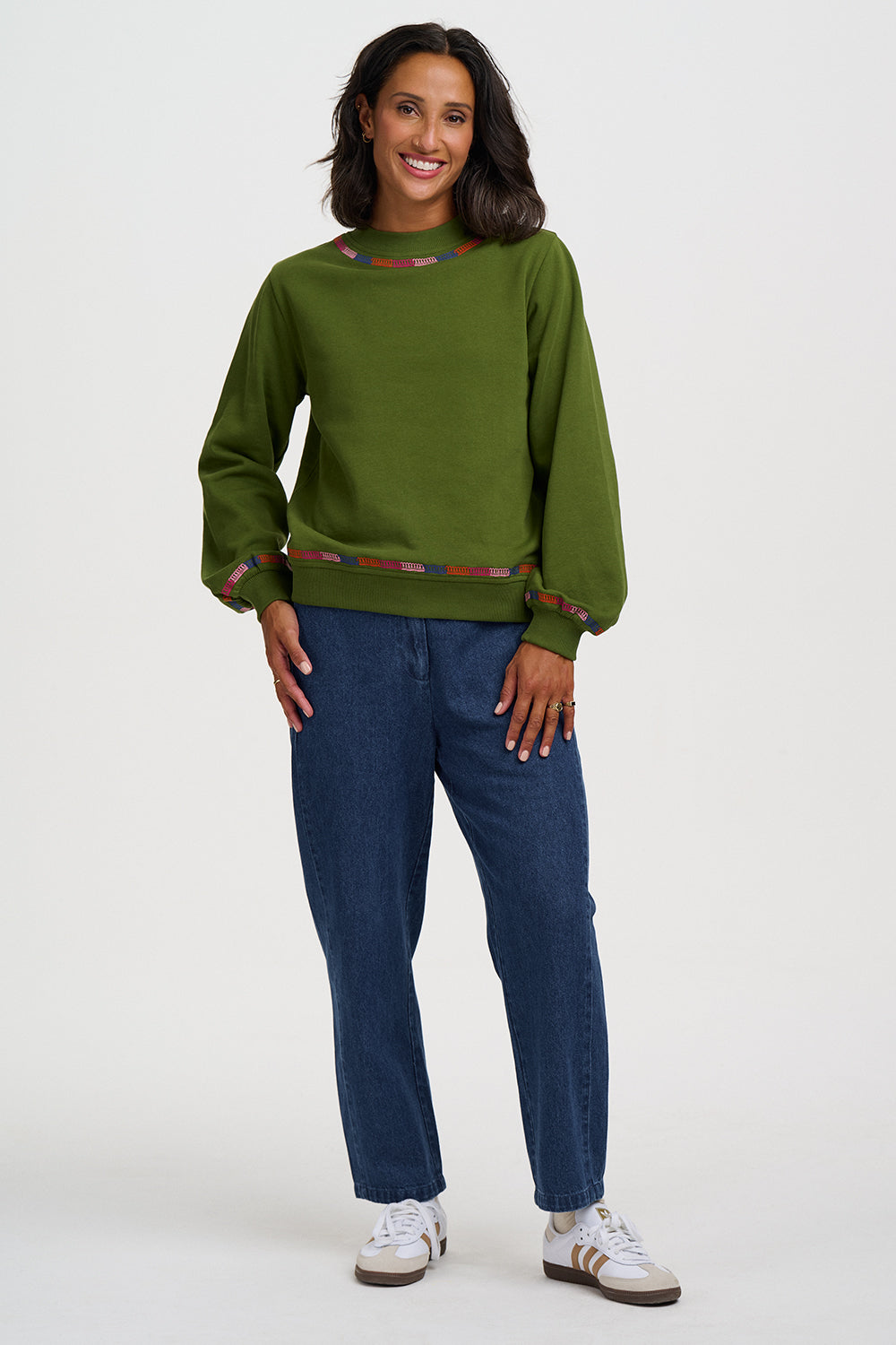 A woman modeling a green long-sleeve crewneck sweatshirt with patterned trim, paired with dark blue jeans and white sneakers, against a light gray studio background.