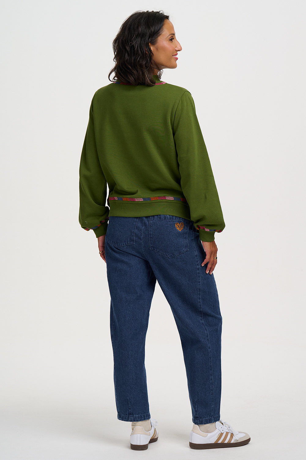 A woman is shown from the back wearing an olive green crewneck sweatshirt, blue jeans, and white sneakers with brown stripes, set against a plain white studio background.
