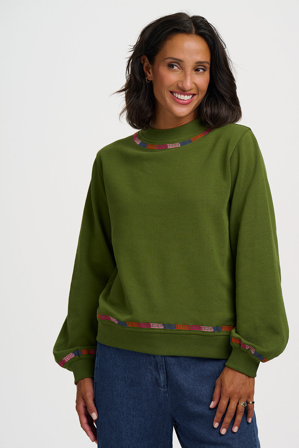 A woman wearing an olive green crewneck sweatshirt with colorful patterned trim at the collar and cuffs, paired with blue jeans, against a light gray studio background.
