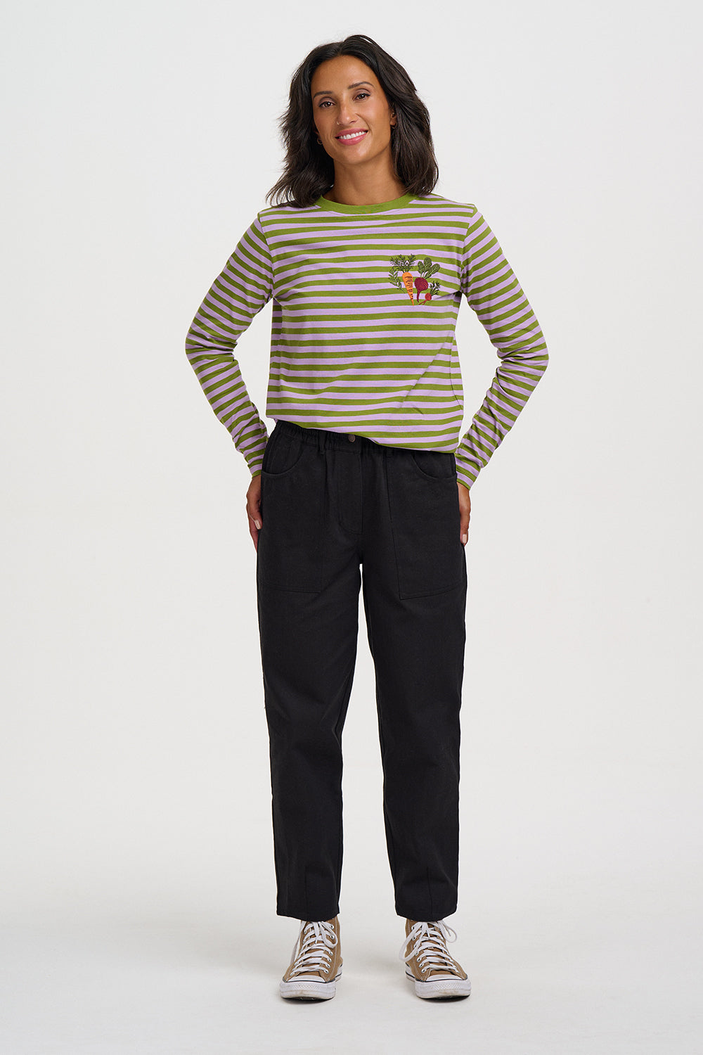A woman modeling a long-sleeve striped shirt with olive green and beige horizontal stripes, featuring a small embroidered vegetable patch on the left chest. She is wearing black pants and beige-and-white sneakers, standing against a plain light gray studio background.