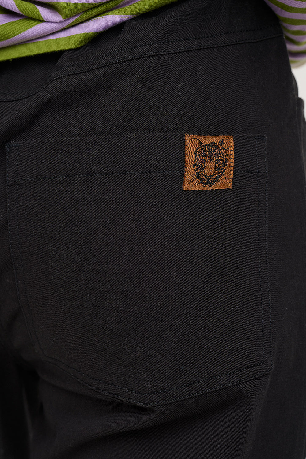 Close-up of a black denim or canvas back pocket with a small brown leather patch featuring a tiger head logo, with a striped shirt edge visible at the top.