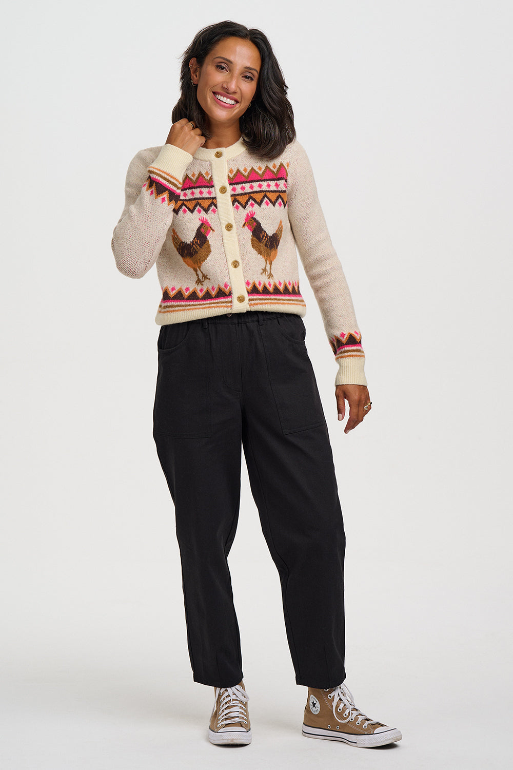 A woman modeling a cream cardigan sweater with a colorful geometric pattern and two brown chickens on the front, paired with black pants and brown sneakers, against a plain light gray background.