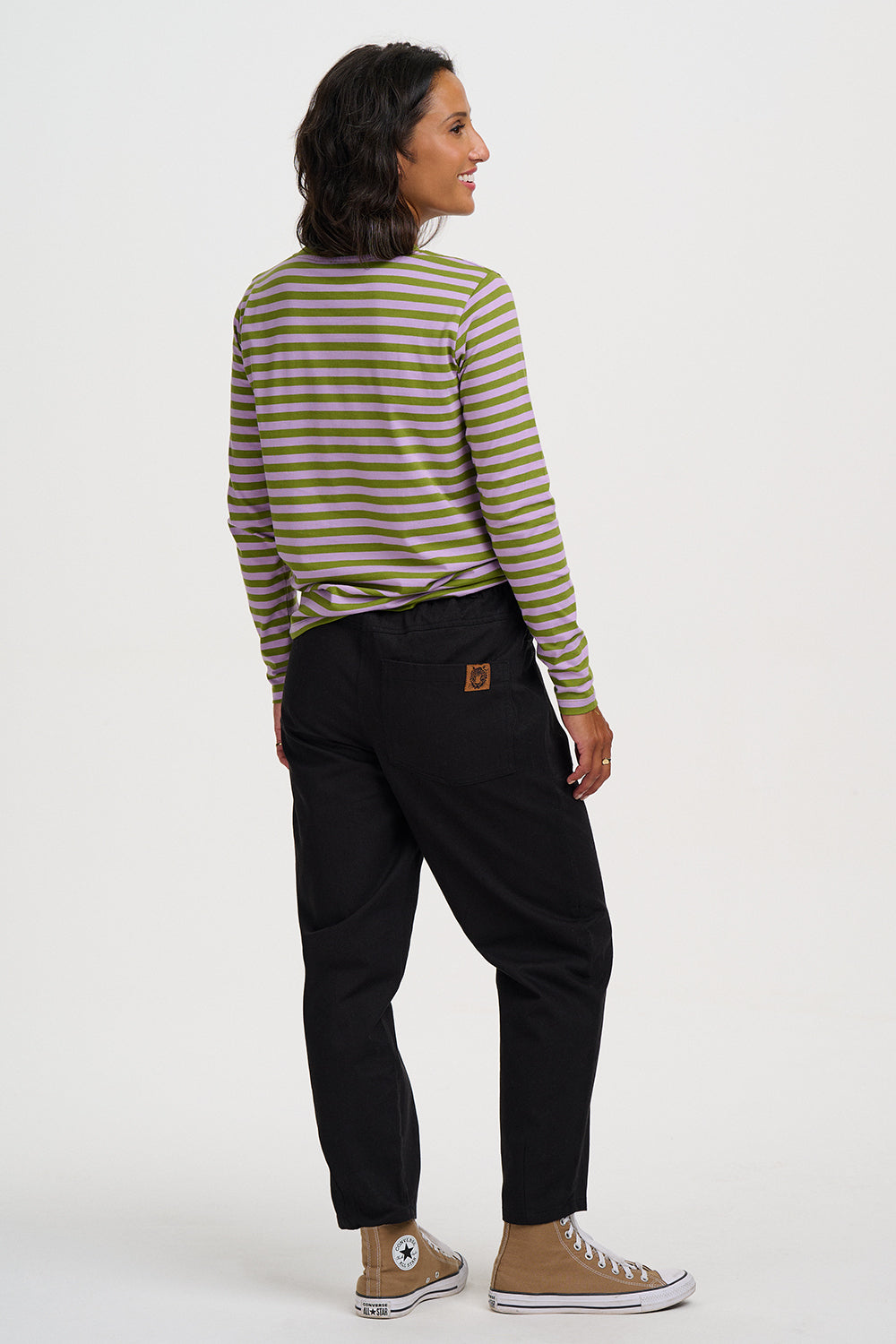 A woman is shown from the back wearing a green and beige striped long-sleeve shirt, black pants, and tan high-top sneakers against a plain white studio background.