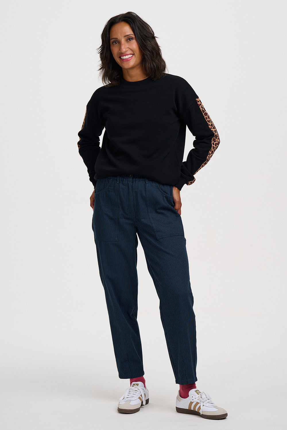 A woman modeling a black crewneck sweatshirt with patterned sleeve detailing, paired with dark navy pants and white sneakers, against a plain light gray studio background.