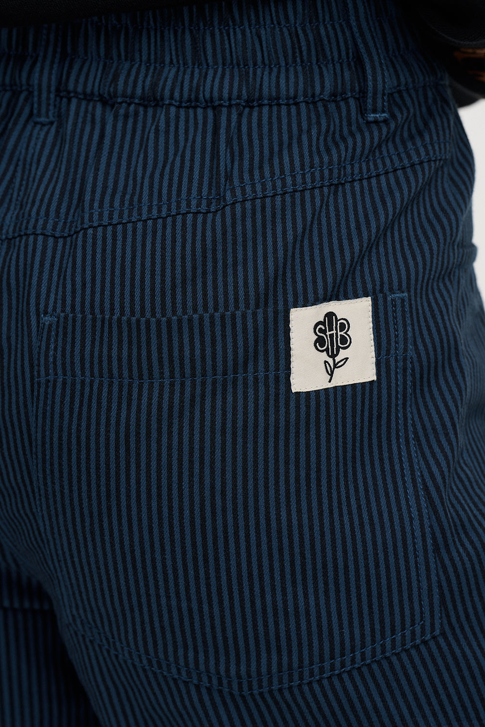 Close-up of dark navy blue pinstriped trousers with a white rectangular patch featuring a black monogram logo on the back pocket.