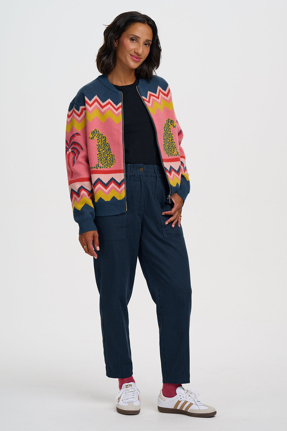 A woman modeling a colorful knit cardigan with pink, yellow, and blue patterns, paired with navy blue pants, a black top, and white sneakers with red socks, against a light gray studio background.