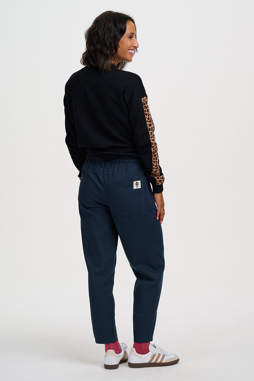 A woman shown from the back wearing a black long-sleeve top with leopard-print sleeve detailing, navy blue pants, and white sneakers with red socks, against a plain light gray studio background.