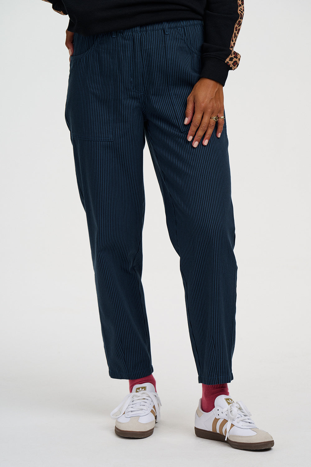 A person wearing dark navy pinstriped trousers, styled with a black top, red socks, and white sneakers with gold stripes, shown from the waist down on a light gray studio background.