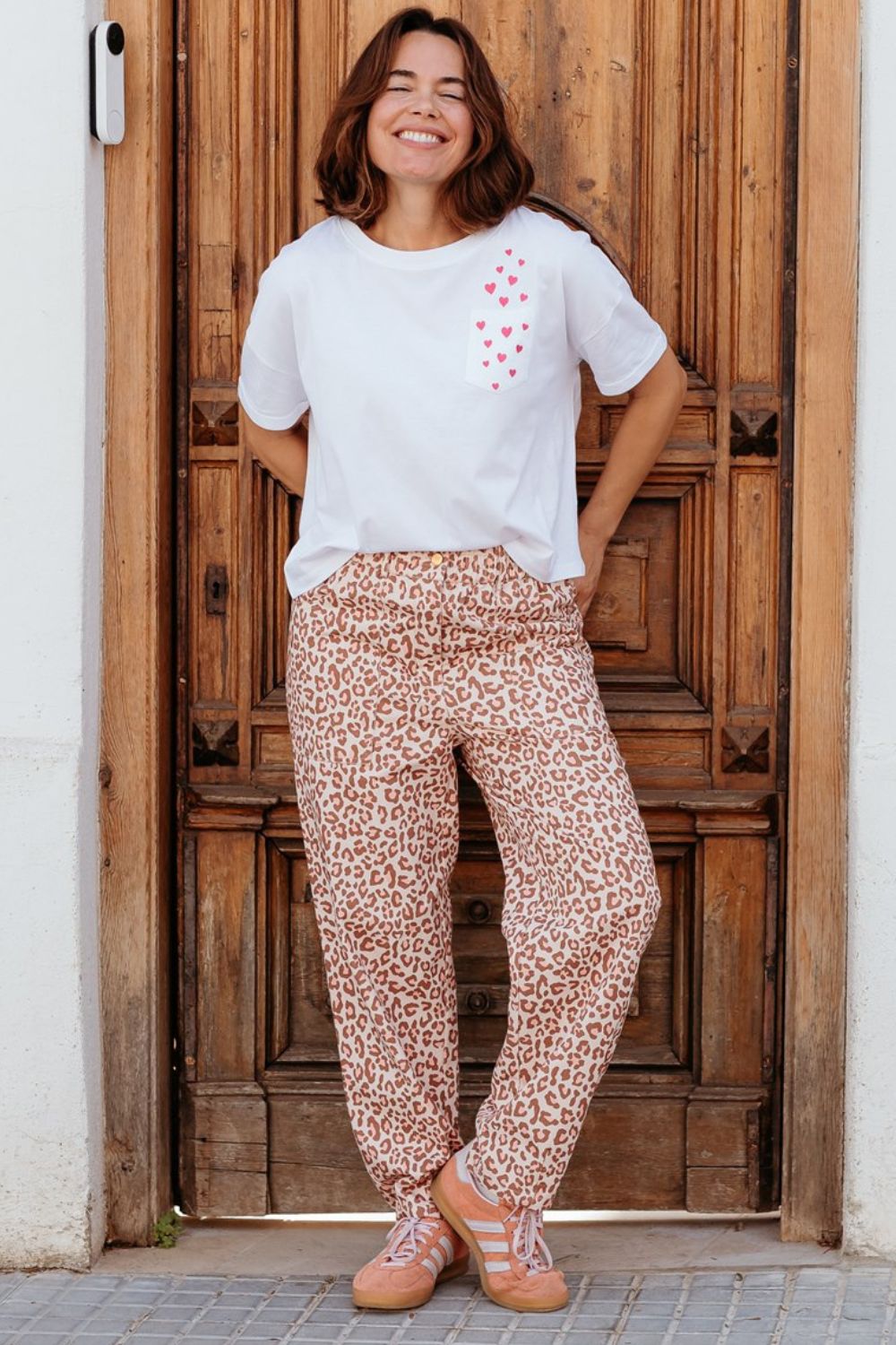 Woman wearing a white t-shirt with a red heart design and patterned pants standing in front of a wooden door.