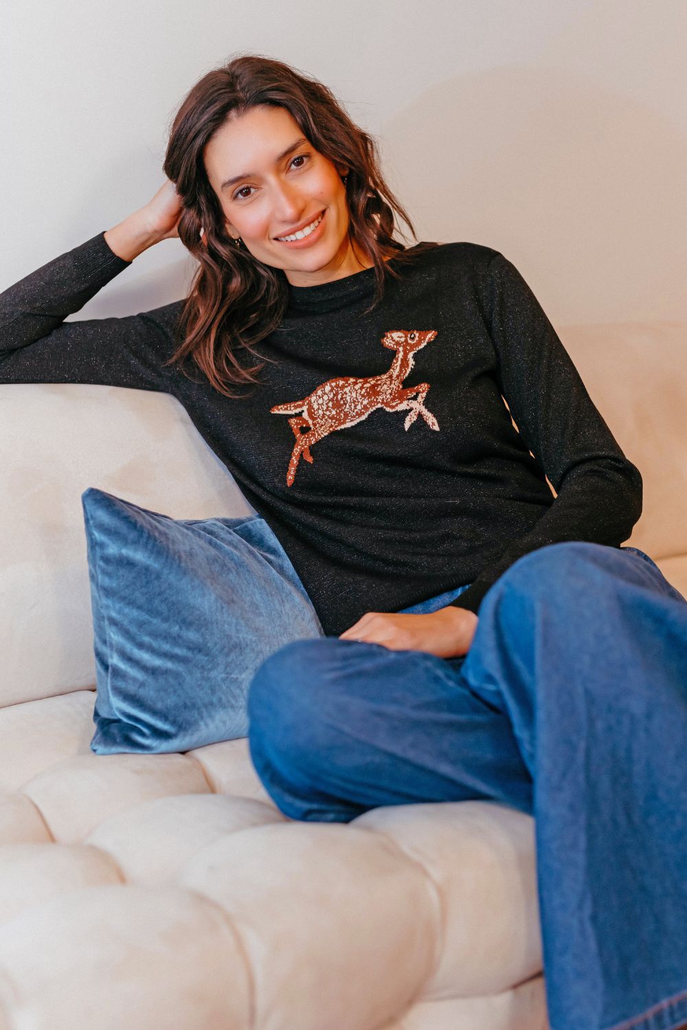 Woman wearing a black sweater with a deer design, sitting on a beige couch with blue pants.