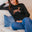 Woman wearing a black sweater with a deer design, sitting on a beige couch with blue pants.