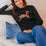 Woman wearing a black sweater with a deer design, sitting on a beige couch with blue pants.
