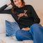 Woman wearing a black sweater with a deer design, sitting on a beige couch with blue pants.