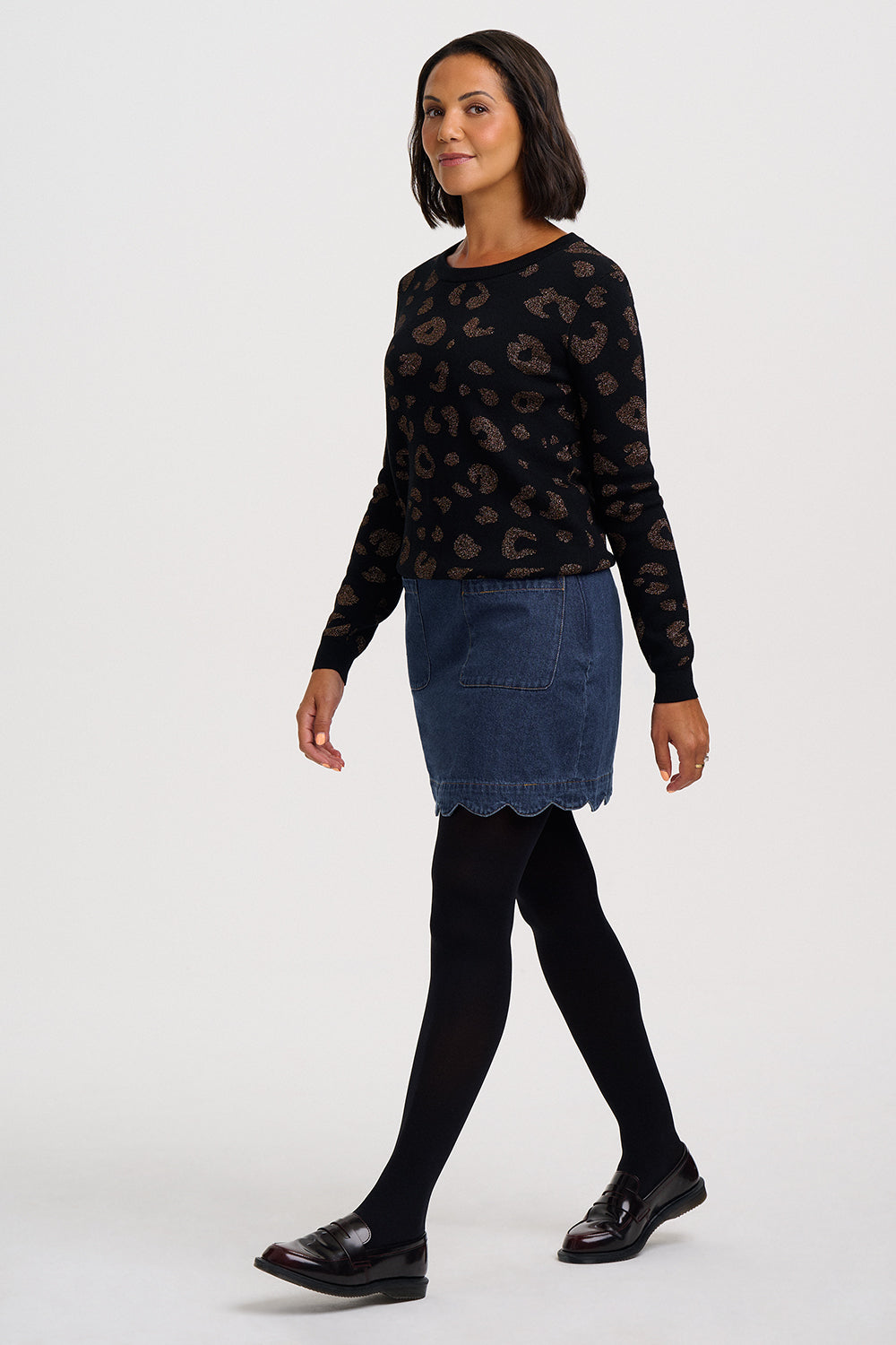 A woman modeling a black long-sleeve knit top with a brown leopard print, paired with a blue denim mini skirt, black tights, and black leather loafers, against a plain light gray studio background.