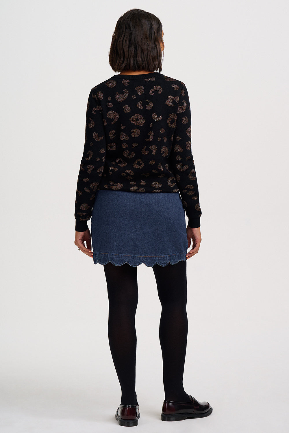 A model shown from the back wearing a black long-sleeve top with a brown paisley pattern, a blue denim mini skirt with a scalloped hem, black tights, and dark brown leather shoes, against a plain white studio background.