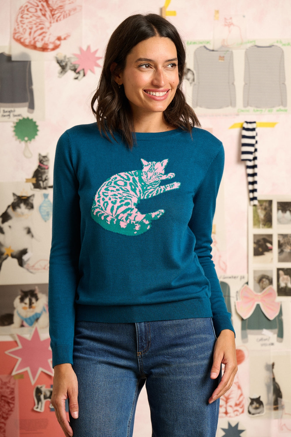 Woman wearing a teal sweater with a cat design in front of a wall with cat-themed decorations.