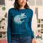 Woman wearing a teal sweater with a cat design in front of a wall with cat-themed decorations.