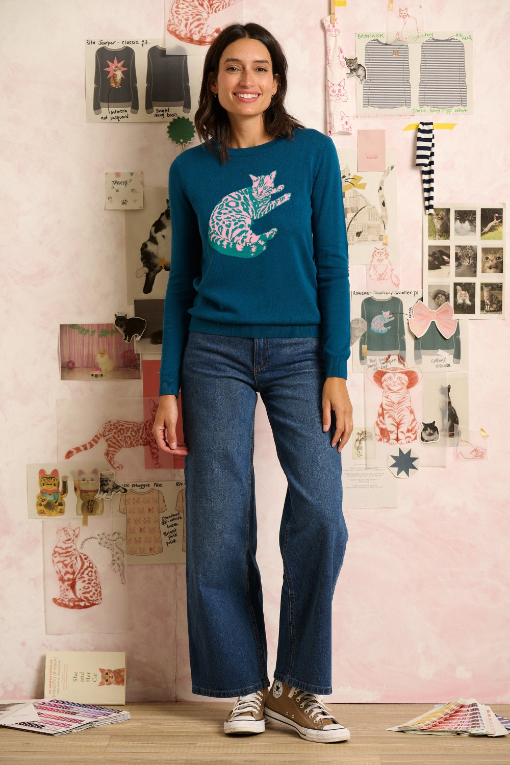 Woman wearing a teal sweater with a colorful cat design and blue jeans, standing in front of a wall with various items.