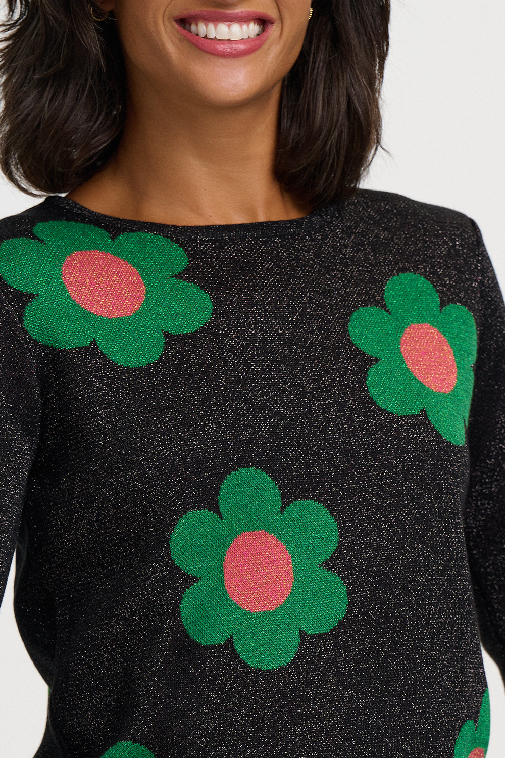 A close-up of a woman wearing a black knit sweater with large green flower motifs and pink centers.