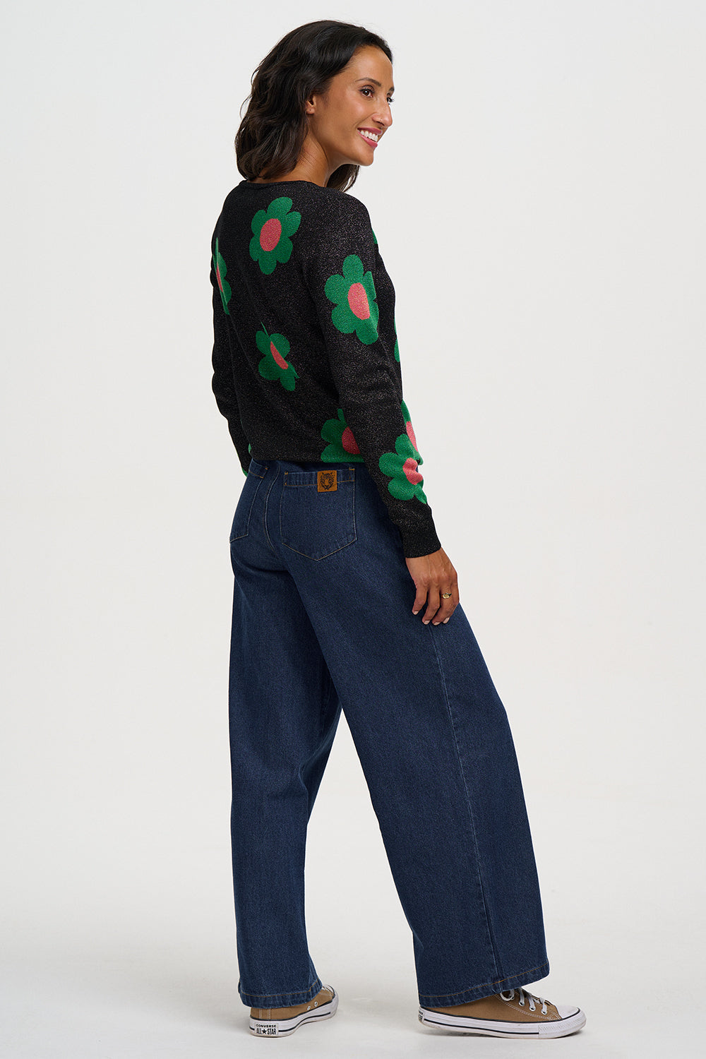 A woman modeling a black knit sweater with large green and pink flower motifs, paired with dark blue wide-leg jeans and brown sneakers, photographed against a plain light gray studio background.