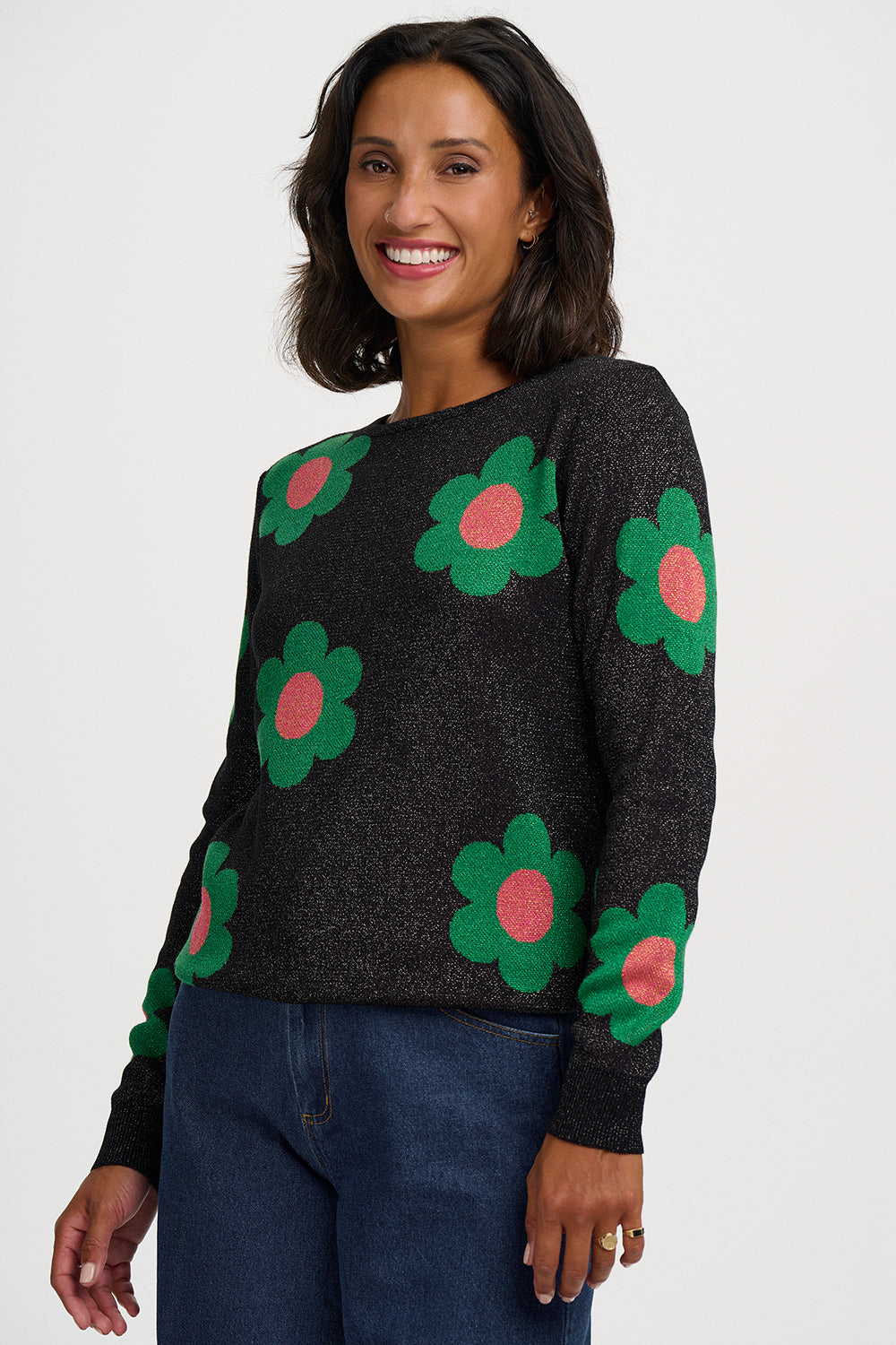 A woman wearing a dark charcoal knit sweater with large green and pink flower motifs, paired with dark blue jeans, against a plain light gray studio background.