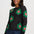 A woman wearing a dark charcoal knit sweater with large green and pink flower motifs, paired with dark blue jeans, against a plain light gray studio background.
