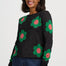 A woman wearing a dark charcoal knit sweater with large green and pink flower motifs, paired with dark blue jeans, against a plain light gray studio background.
