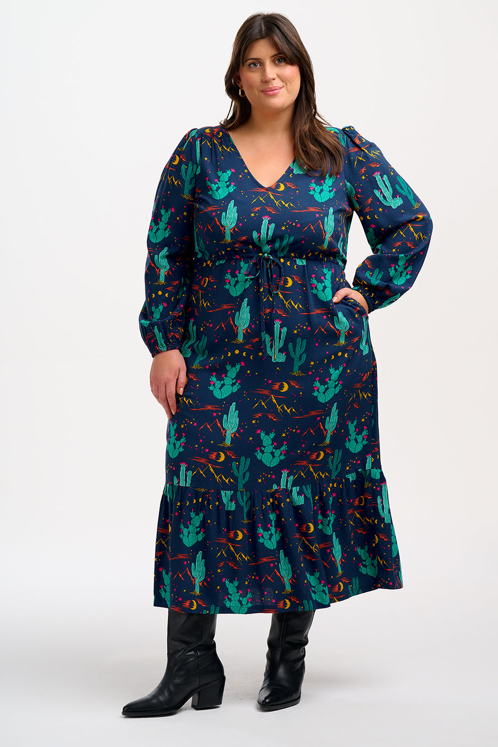 A woman standing and posing in a navy blue maxi dress with a colorful desert pattern, featuring V-neck, long blouson sleeves, a drawstring waistband, and a tiered hem.