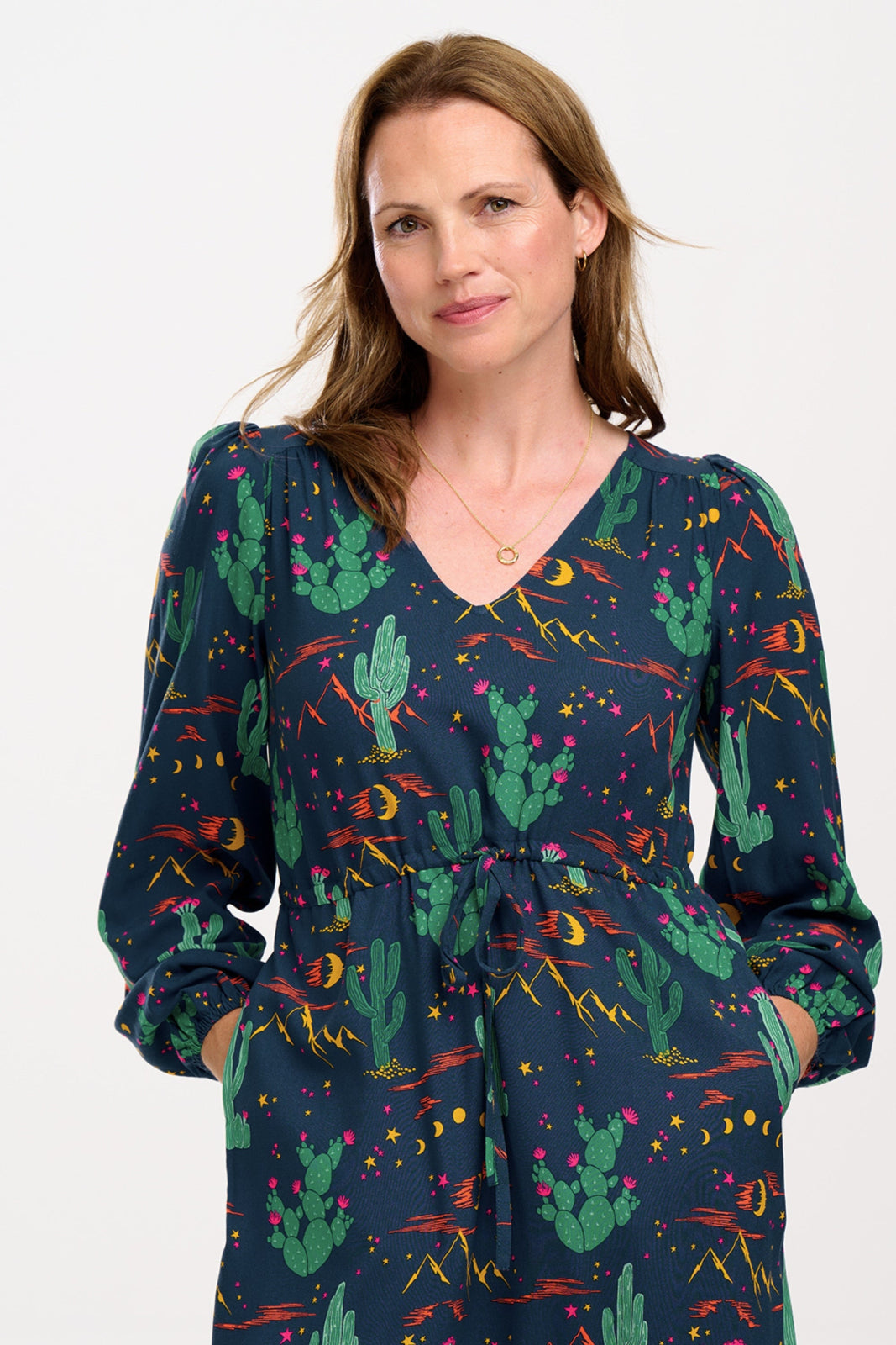 A woman standing and posing in a navy blue maxi dress with a colorful desert pattern, featuring V-neck, long blouson sleeves, a drawstring waistband, and a tiered hem.