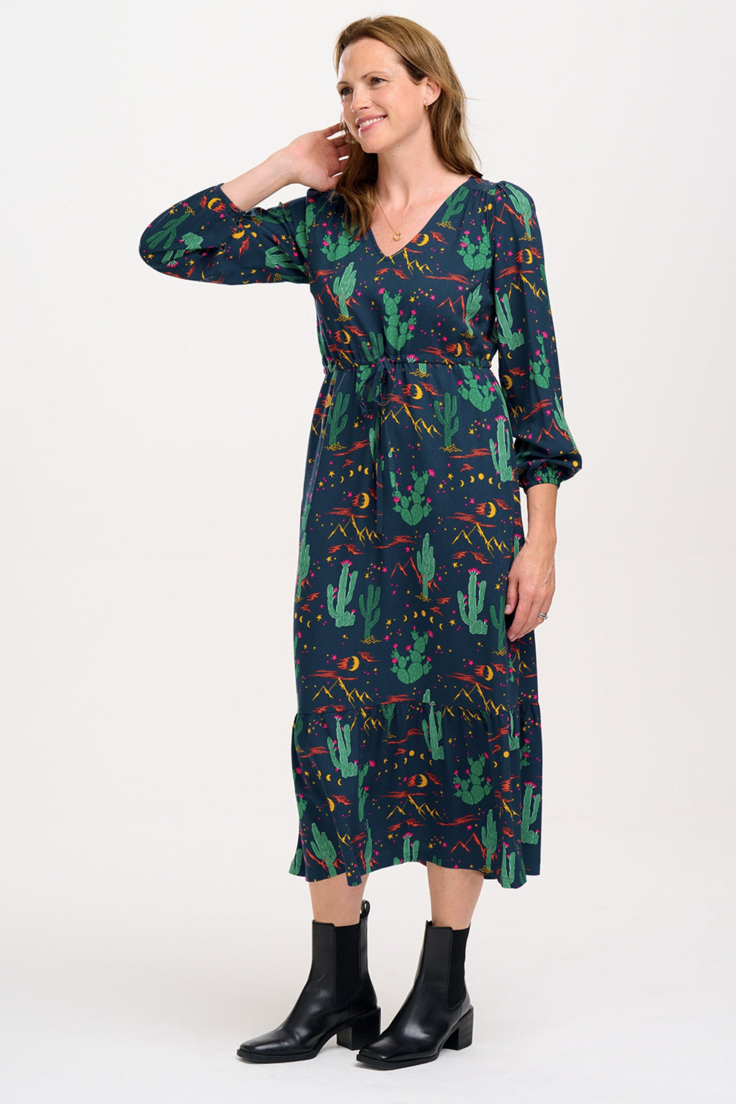 A woman standing and posing in a navy blue maxi dress with a colorful desert pattern, featuring V-neck, long blouson sleeves, a drawstring waistband, and a tiered hem.