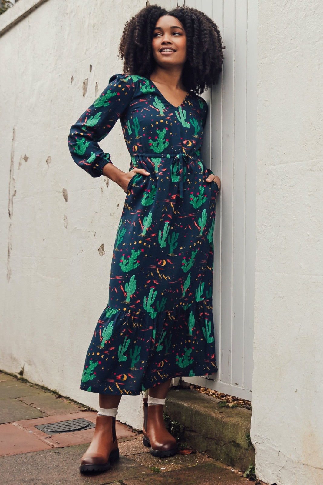 A woman standing against a wall, wearing a navy-colored maxi dress with a geometric pattern, featuring a V-neck, long sleeves, a drawstring waistband, and a tiered hem.