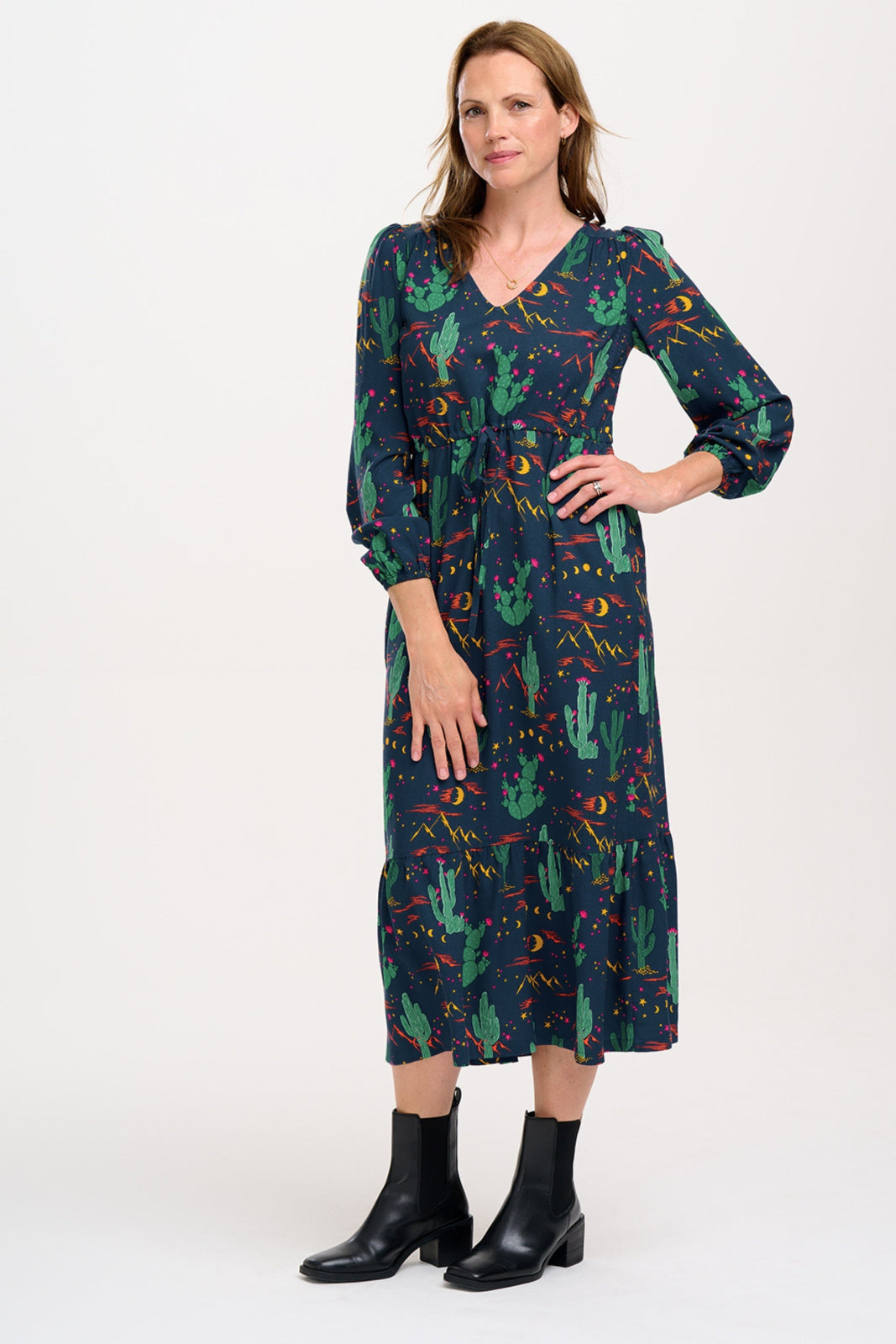 A woman standing and posing in a navy blue maxi dress with a colorful desert pattern, featuring V-neck, long blouson sleeves, a drawstring waistband, and a tiered hem.