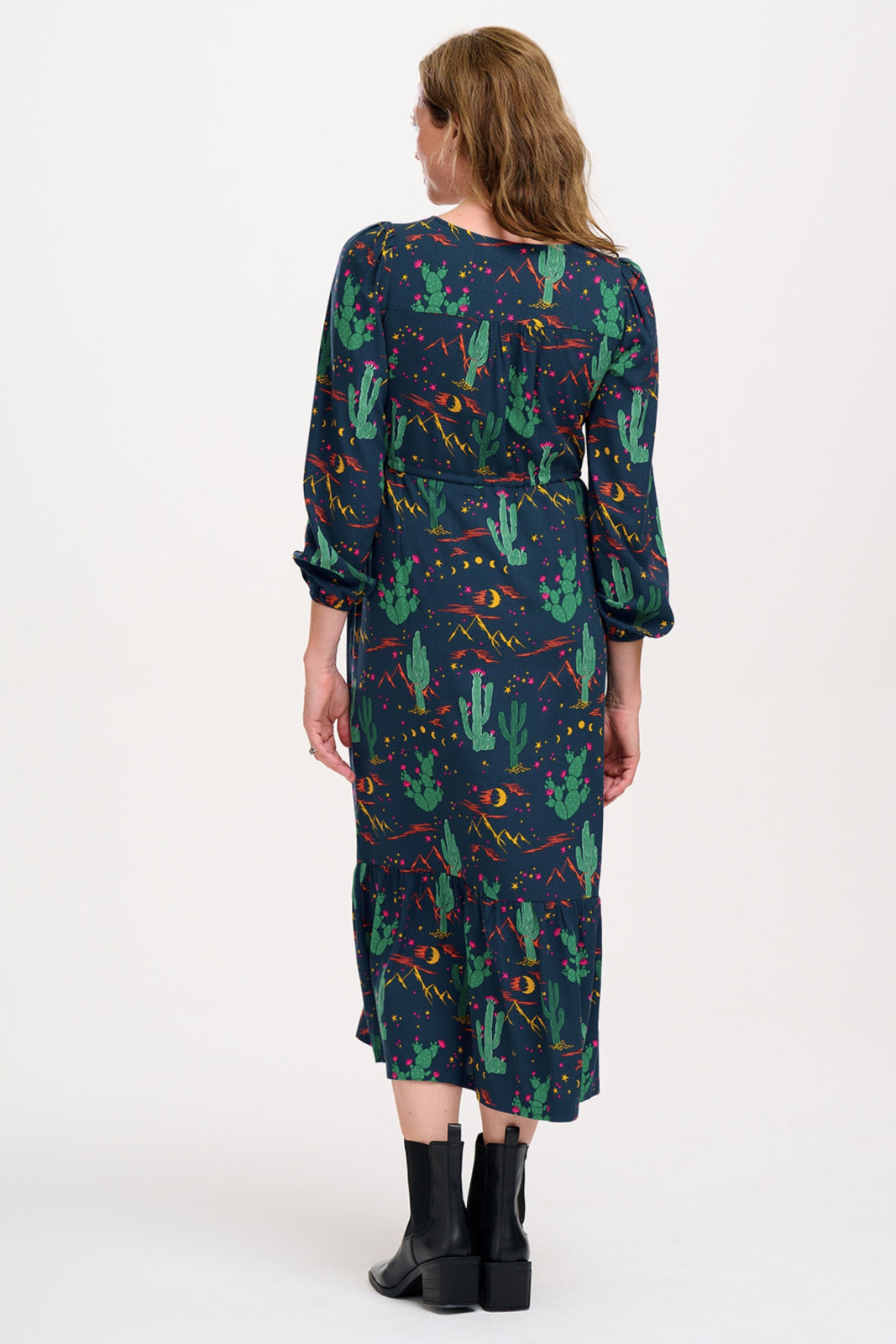 A woman standing and posing in a navy blue maxi dress with a colorful desert pattern, featuring V-neck, long blouson sleeves, a drawstring waistband, and a tiered hem.