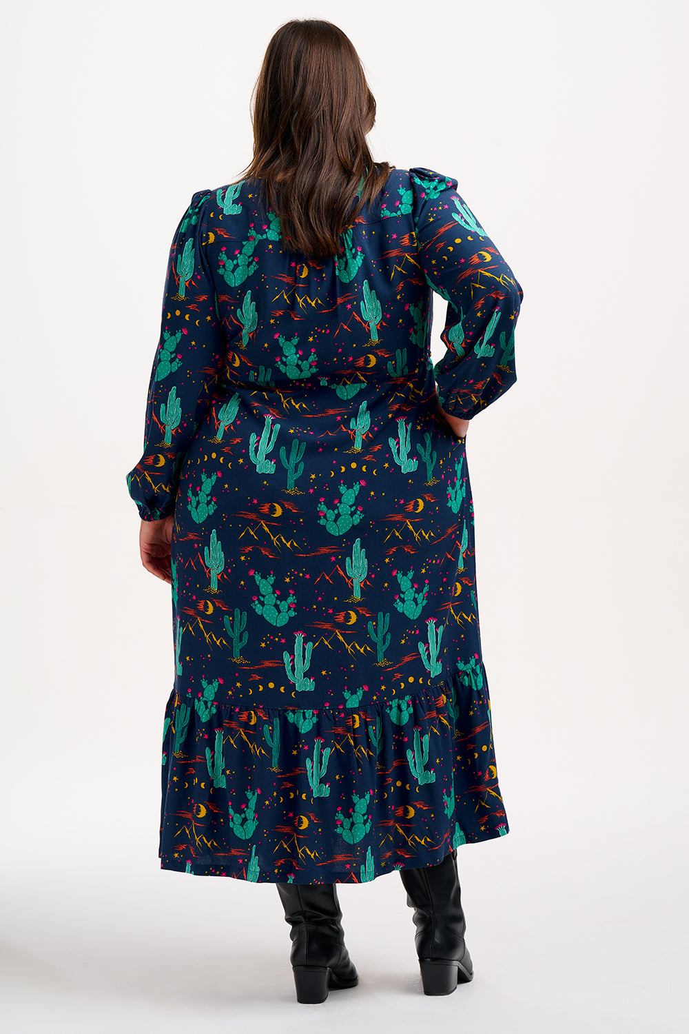 A woman standing and posing in a navy blue maxi dress with a colorful desert pattern, featuring V-neck, long blouson sleeves, a drawstring waistband, and a tiered hem.