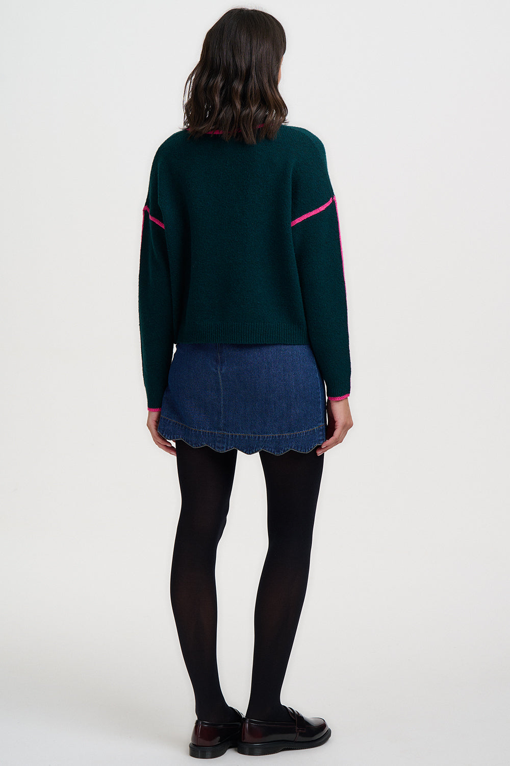 Person wearing a dark green sweater with pink accents and a denim skirt on a white background