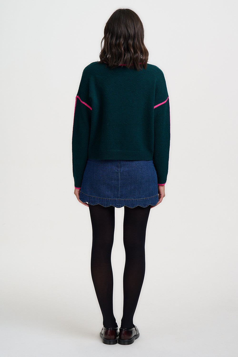 Person wearing a dark green sweater with pink accents and a denim skirt on a white background