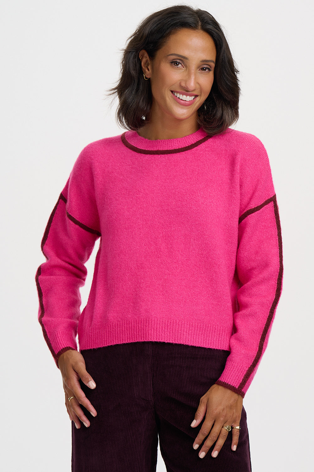A woman wearing a bright pink knit sweater with dark trim on the crew neck, cuffs, hem, and along the sleeves, paired with dark burgundy pants, against a light gray studio background.