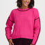 A woman wearing a bright pink knit sweater with dark trim on the crew neck, cuffs, hem, and along the sleeves, paired with dark burgundy pants, against a light gray studio background.