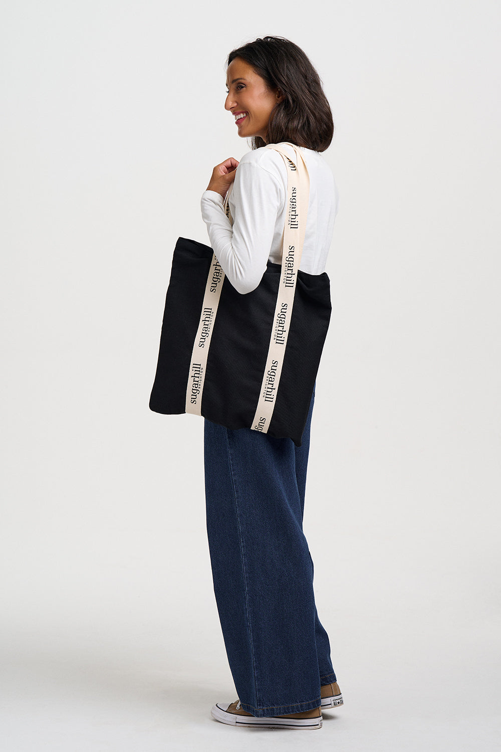 A woman wearing a white long-sleeve shirt, wide-leg blue jeans, and brown sneakers is shown from the back, carrying a black canvas tote bag with cream-colored shoulder straps printed with the brand name 'BAGS OF LOVE'.