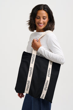 A woman wearing a white long-sleeve top and blue jeans carries a large black tote bag with cream-colored shoulder straps printed with the brand name 'sugarchill'.