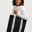 A woman wearing a white long-sleeve top and blue jeans carries a large black tote bag with cream-colored shoulder straps printed with the brand name 'sugarchill'.