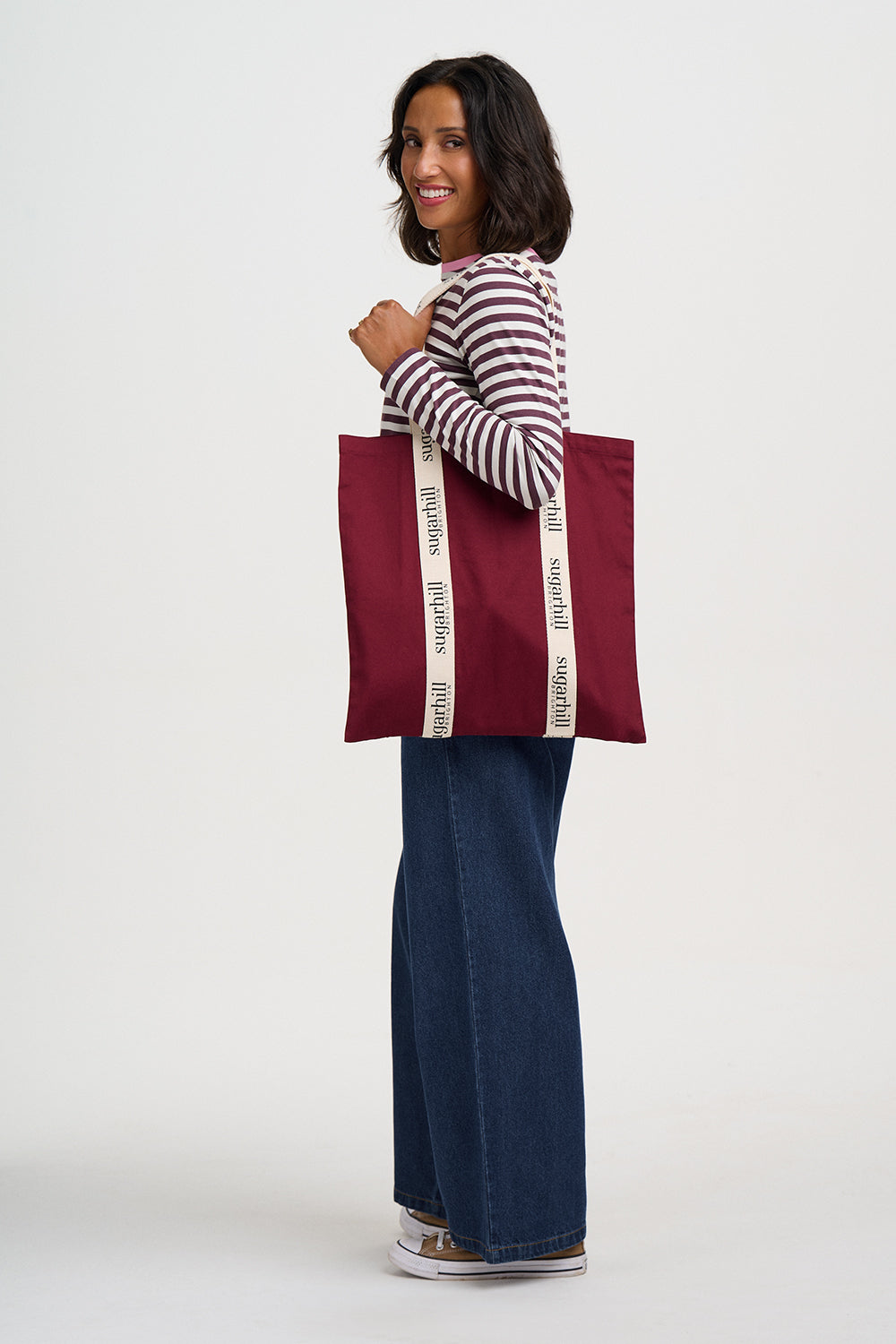 A woman wearing a striped long-sleeve shirt, wide-leg denim jeans, and sneakers is shown from the side, holding a large burgundy tote bag with cream-colored shoulder straps printed with the brand name 'sophia' in black.